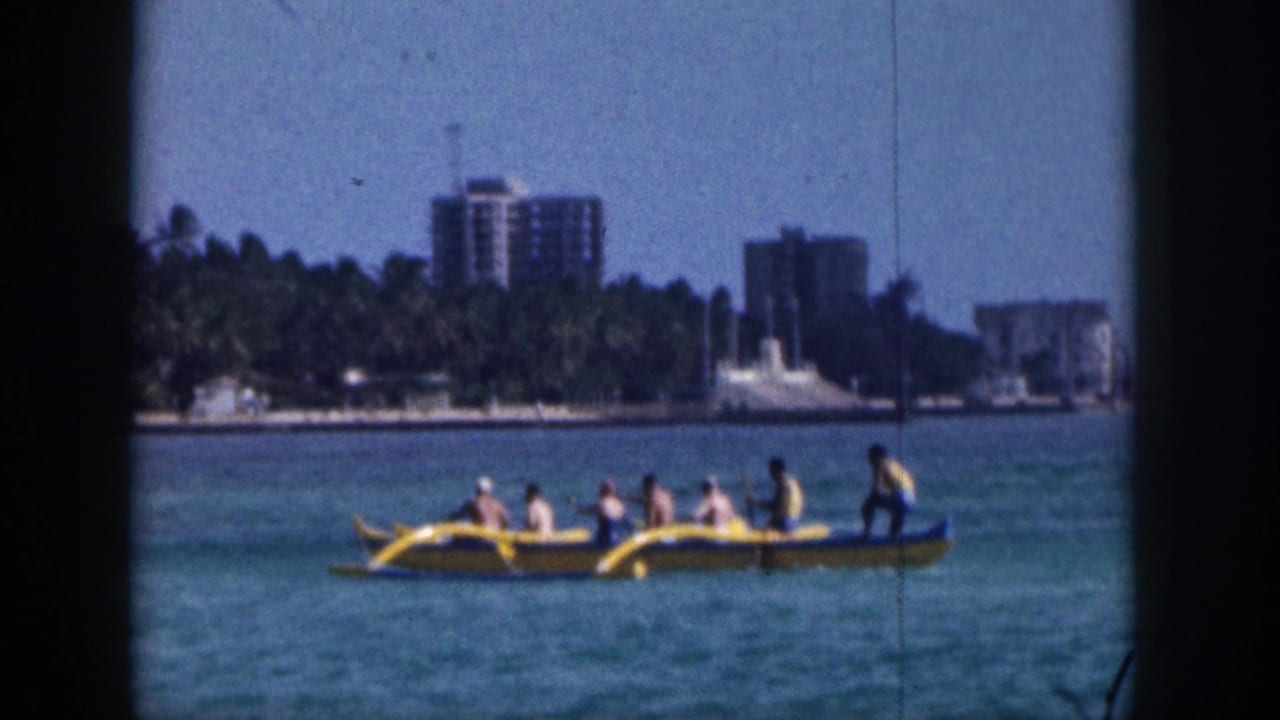 Outrigger canoe with passengers moves across turquoise water in front of shoreline buildings.