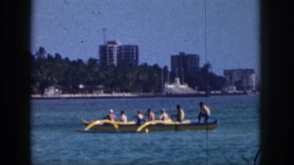 1959 Hawaii home-movie frame of a yellow outrigger canoe carrying a small group across turquoise water near a palm-lined shore.