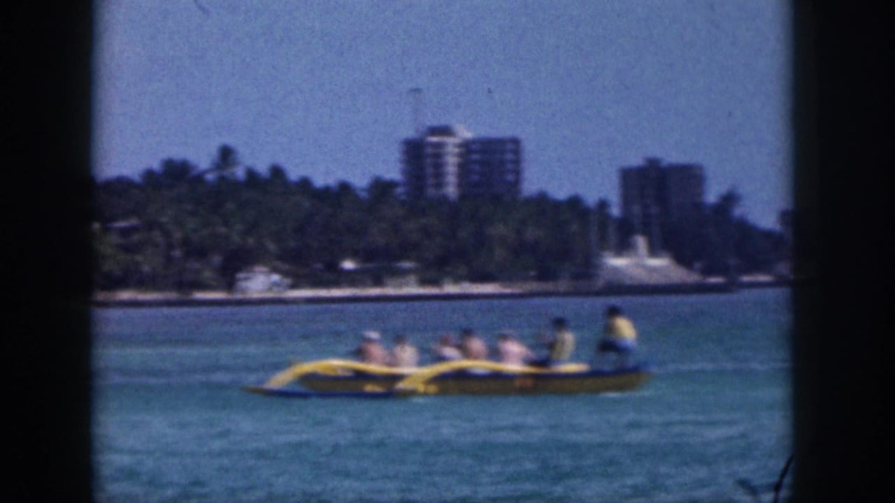 Soft 8mm frame from Hawaii in 1959 showing a yellow outrigger canoe on blue water with palms and waterfront buildings beyond.