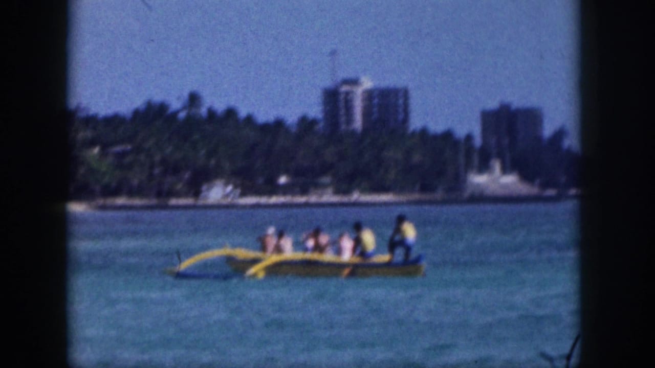 1959 Hawaii frame showing several people in a yellow outrigger canoe on calm water with a palm-lined shoreline and buildings behind them.