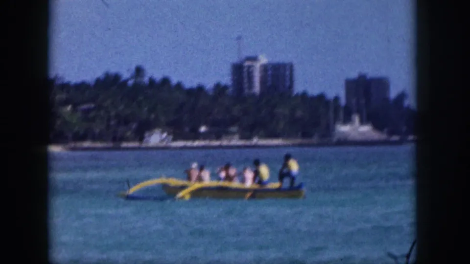 1959 Hawaii frame showing several people in a yellow outrigger canoe on calm water with a palm-lined shoreline and buildings behind them.