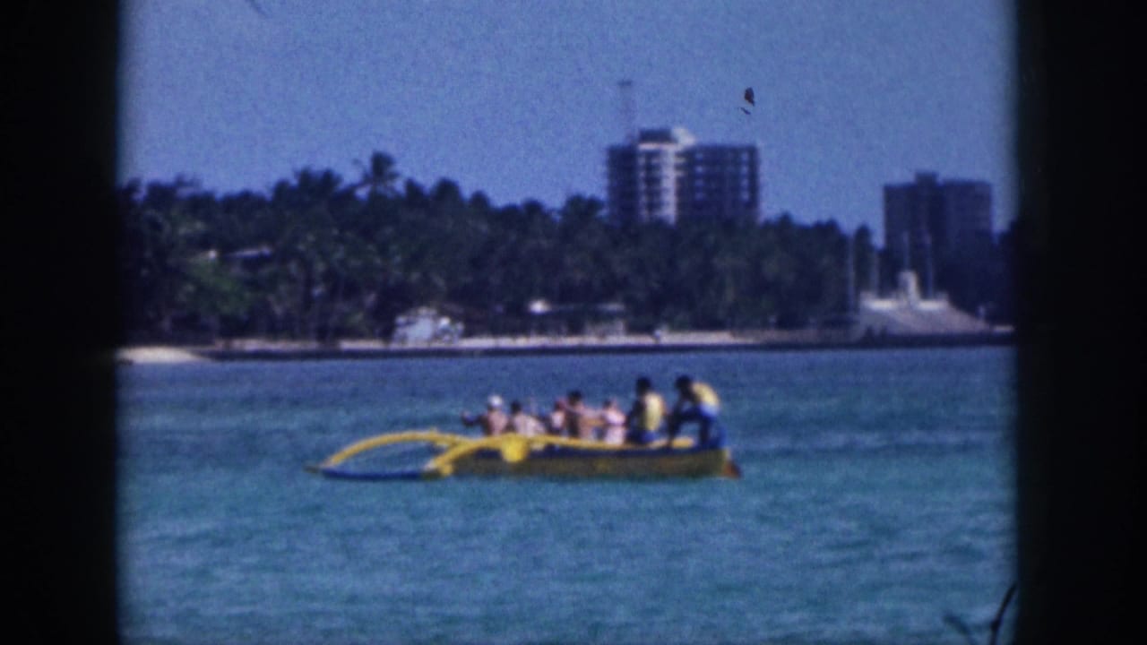 1959 Hawaii home-movie frame showing a yellow outrigger canoe on turquoise water with palms and high-rise buildings in the distance.