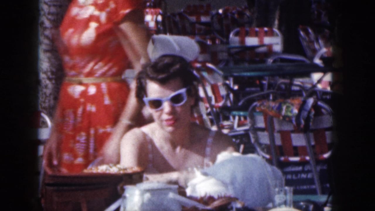 Woman in white sunglasses seated at an outdoor cafe beside a standing woman in a red dress.