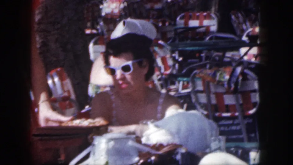 1959 Hawaii home-movie frame of a woman in white sunglasses seated at an outdoor cafe, surrounded by striped chairs and bright resort furnishings.