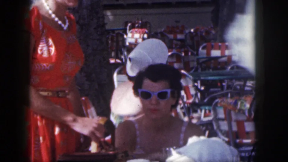 1959 Hawaii resort cafe frame showing a seated woman in sunglasses, a standing woman in a red dress, striped chairs, and table items in bright sun.