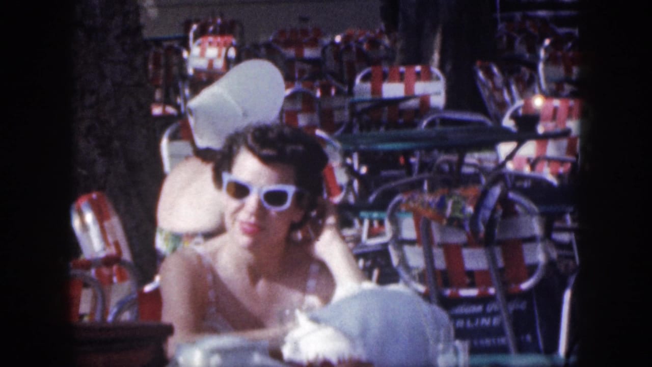 Woman in white sunglasses seated at a beach cafe with red and white striped chairs behind her.