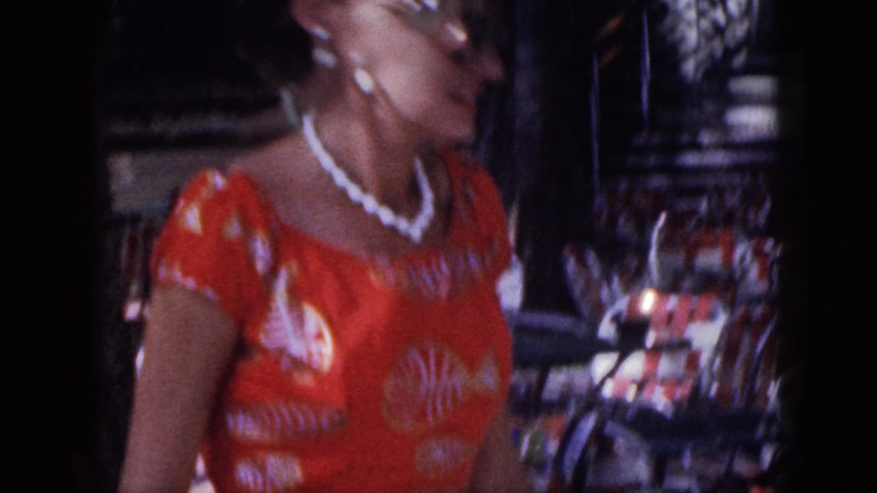 Woman in an orange patterned dress and pearl necklace walking past outdoor cafe seating.