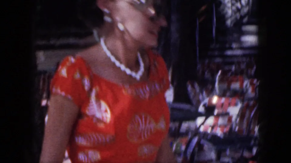 1959 Hawaii home-movie frame of a woman in an orange patterned dress passing in front of beach-cafe chairs.