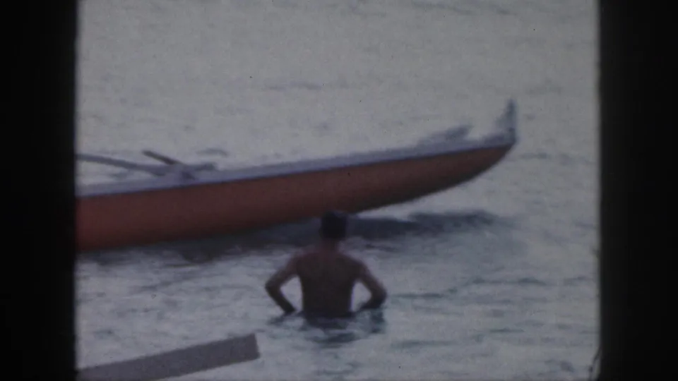 1959 Hawaii home-movie frame of a swimmer near the hull of an outrigger canoe in calm ocean water.