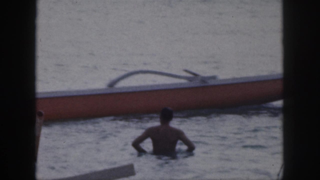 Shirtless man in the water near an outrigger canoe, seen through dark frame edges.