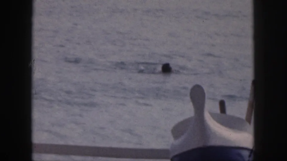 1959 Hawaii home-movie frame with distant swimmers and a small boat bow seen from a waterfront deck.