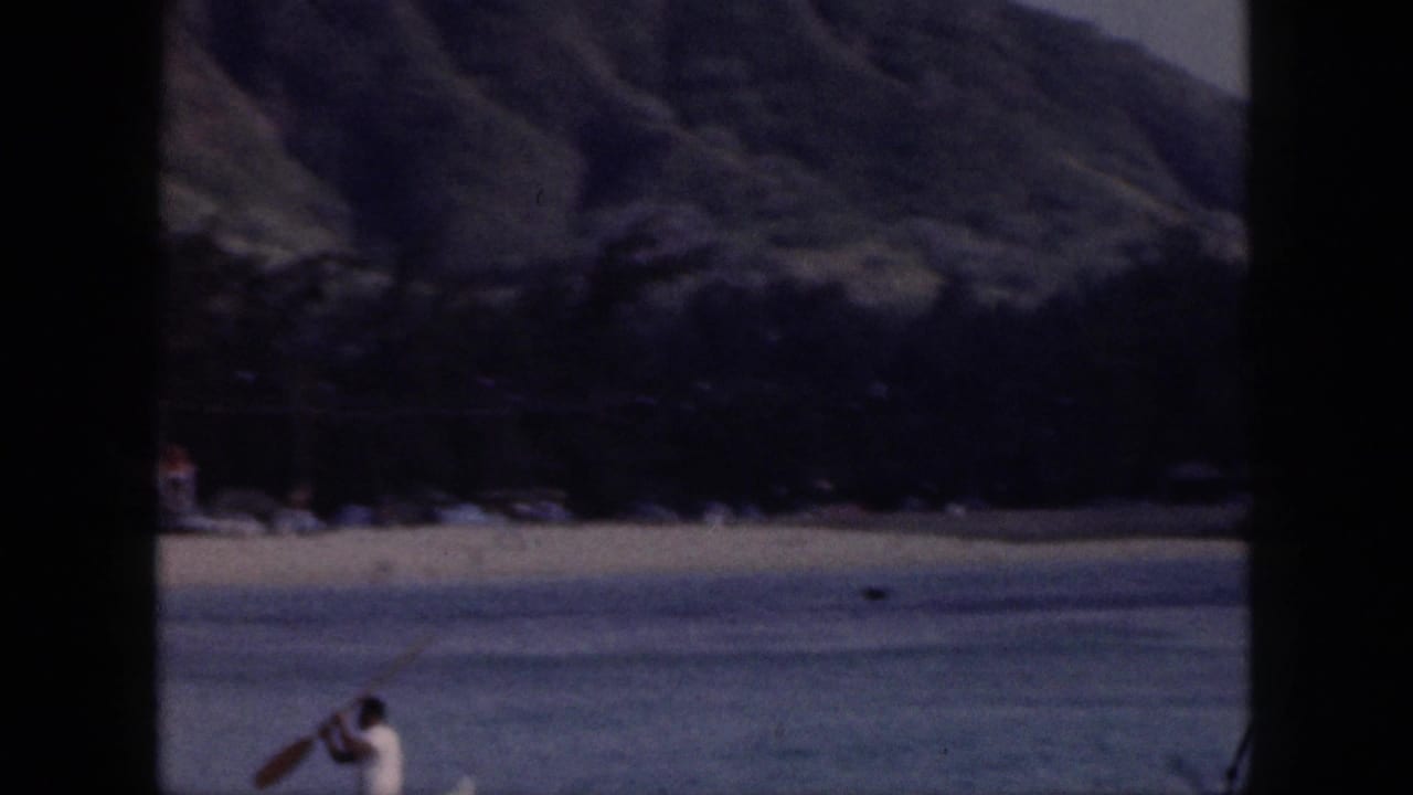 Distant paddler on water near a beach and mountain backdrop.