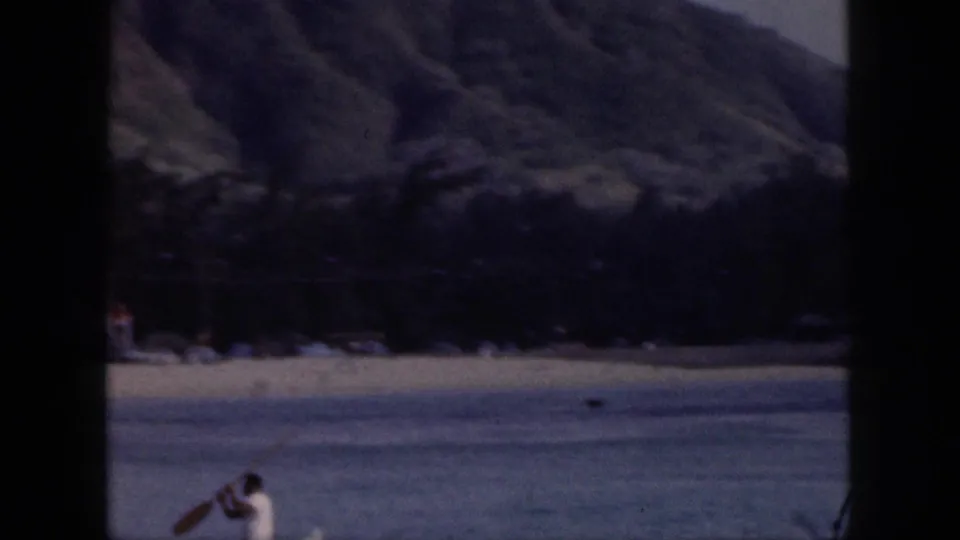 1959 Hawaii 8mm frame with a distant person paddling on the water below a beach, trees, and mountains.
