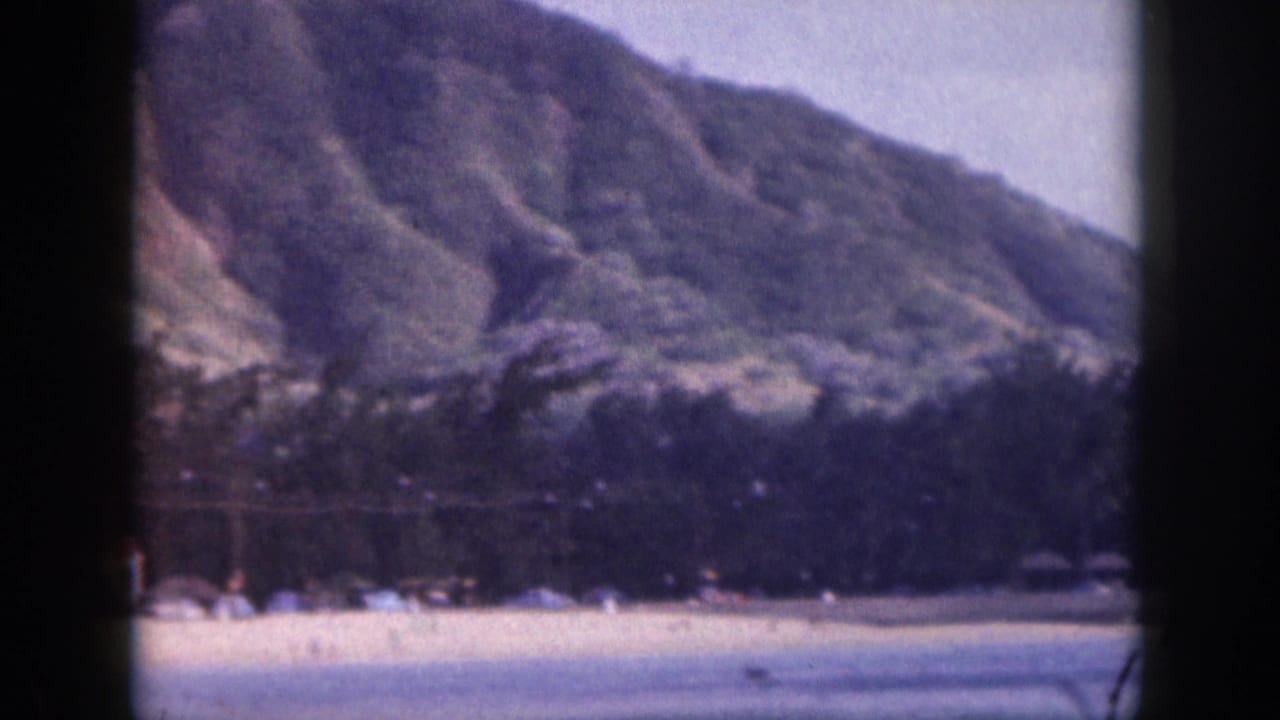 1959 Hawaii 8mm frame showing a pale beach, distant people or beach objects, trees, and steep mountains.