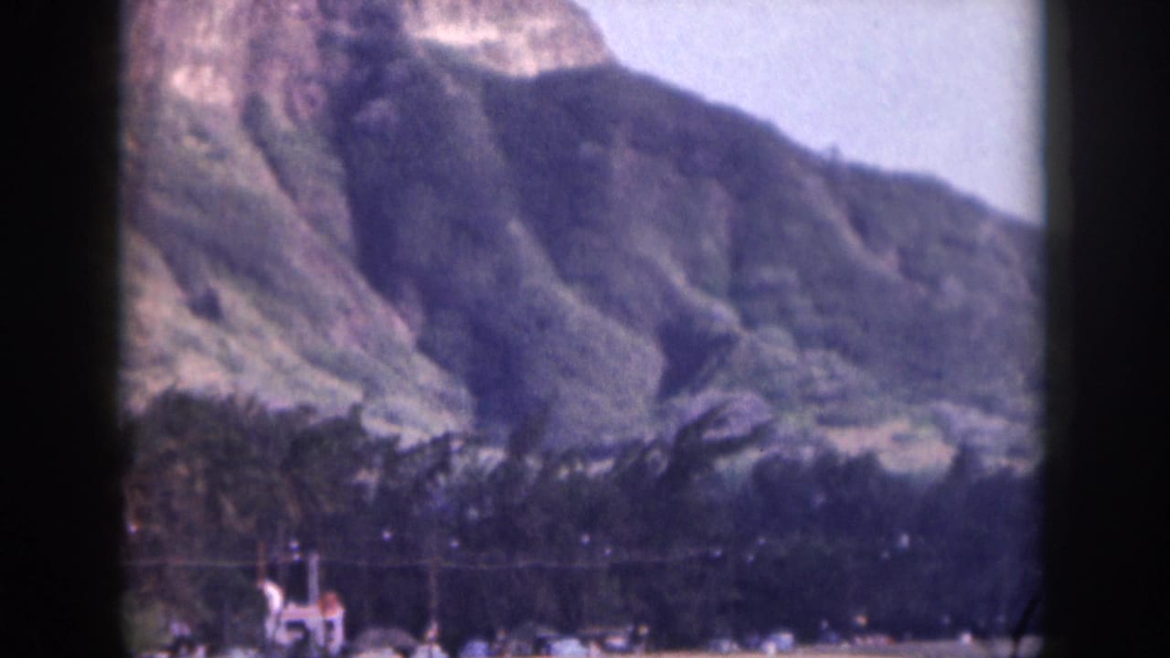 1959 Hawaii 8mm frame with a mountain-backed shoreline, beach activity, and a small craft or swimmers near the water.