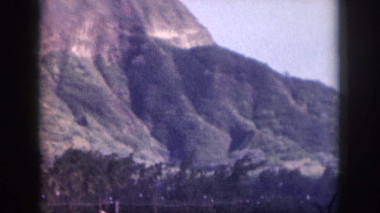 1959 Hawaii 8mm frame showing distant rugged mountain slopes, green lower vegetation, pale sky, and dark gate edges.