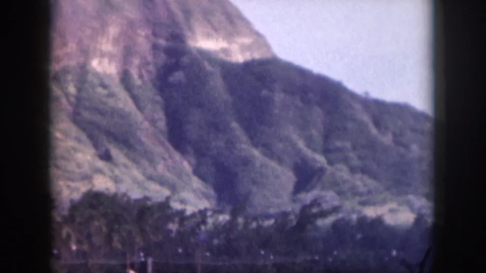 1959 Hawaii 8mm frame showing distant rugged mountain slopes, green lower vegetation, pale sky, and dark gate edges.