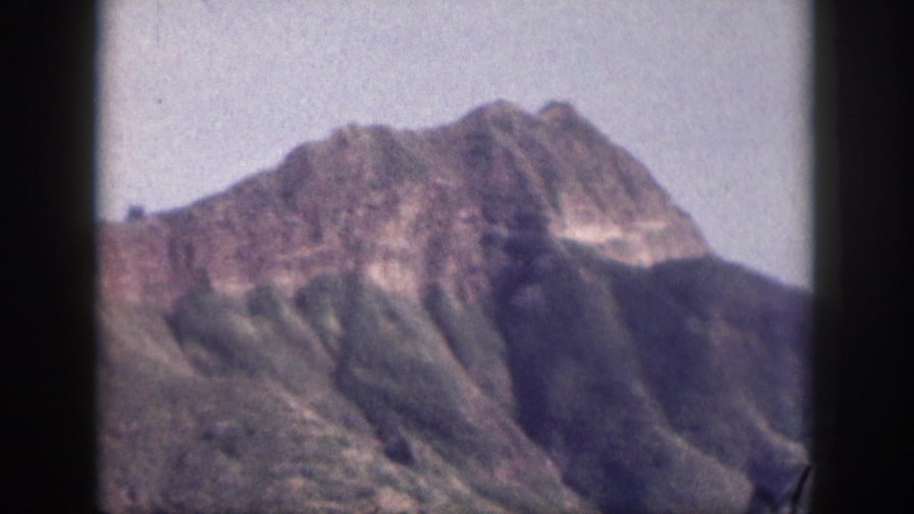 1959 Hawaii 8mm still with a high rocky peak, shadowed green mountainside, pale sky, and softened home-movie focus.