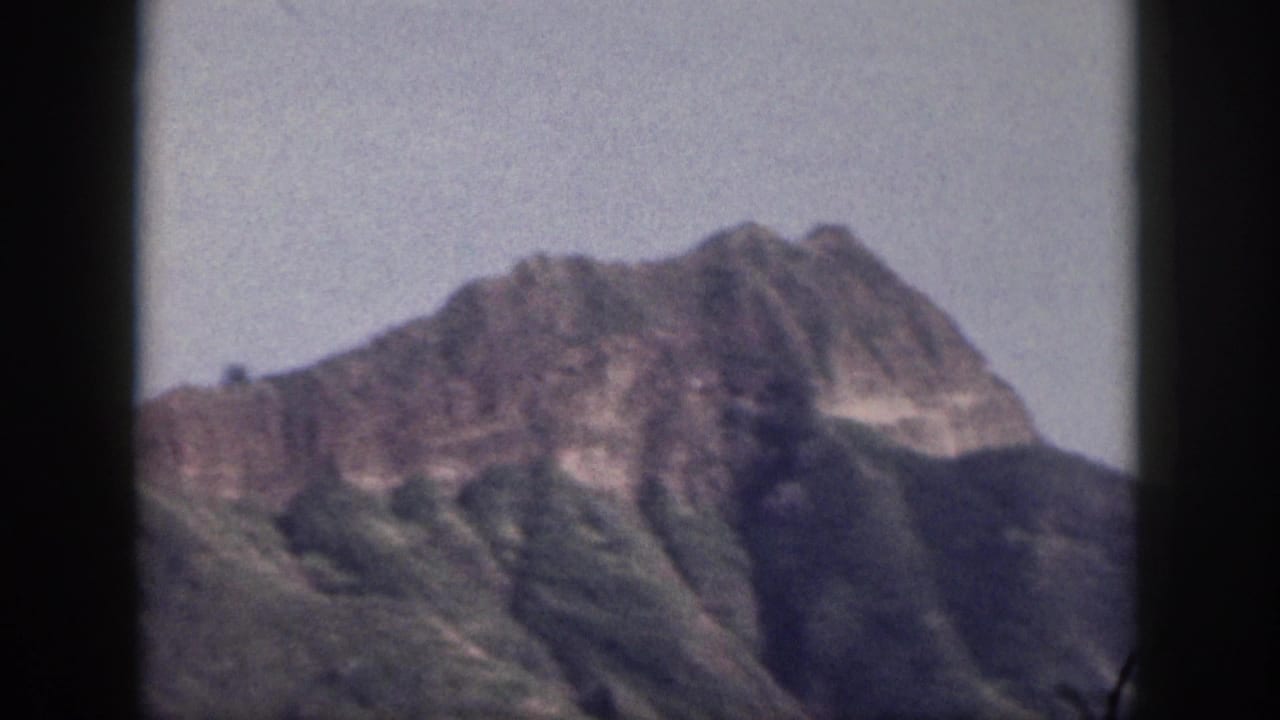 1959 Hawaii home-movie frame showing a distant peak, steep green ridges, pale sky, and dark 8mm gate borders.