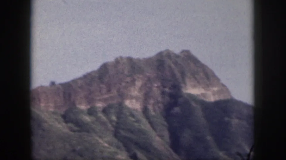 1959 Hawaii home-movie frame showing a distant peak, steep green ridges, pale sky, and dark 8mm gate borders.