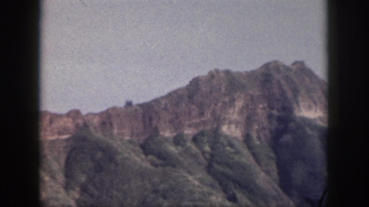 Rocky mountain ridge and green slopes beneath a pale sky, framed by dark film edges.