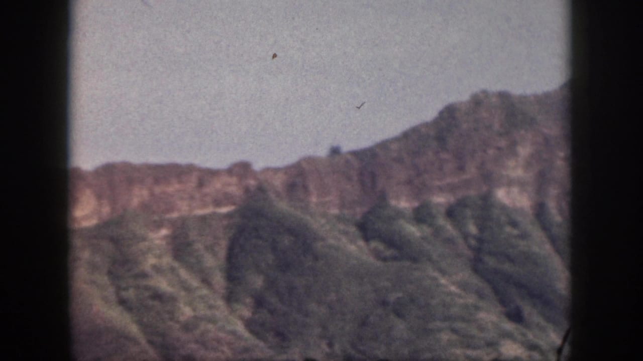 Mountain ridge with shadowed green slopes and pale blue sky, bordered by film frame edges.