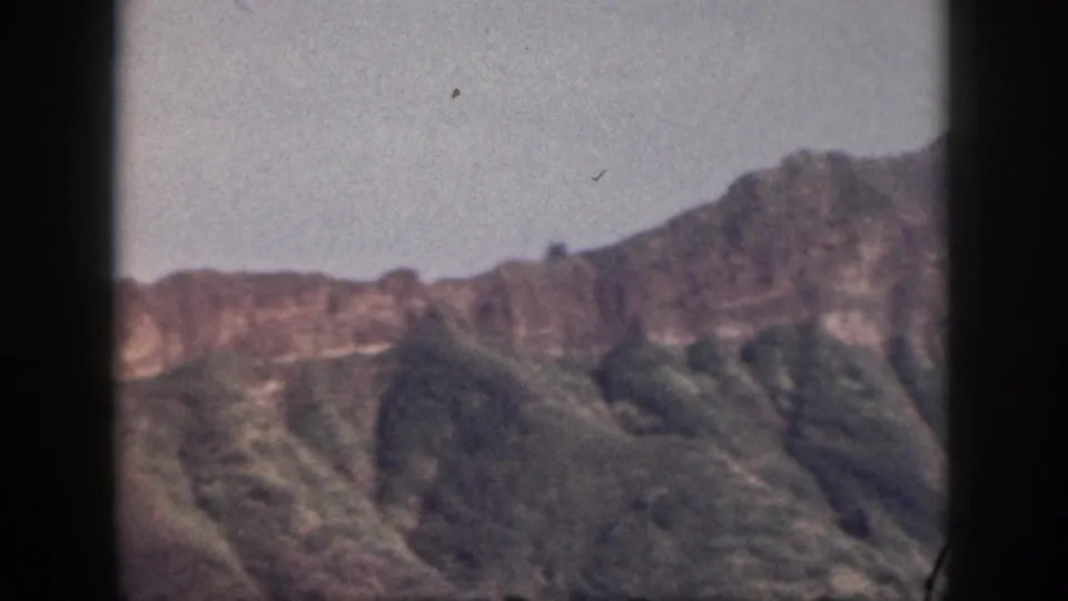 1959 Hawaii home-movie still of a rocky ridge line and shadowed green slopes under a pale blue sky.