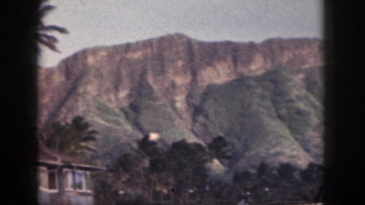 1959 Hawaii coastal view with rugged green mountains, palm trees, and low buildings in soft home-movie color.