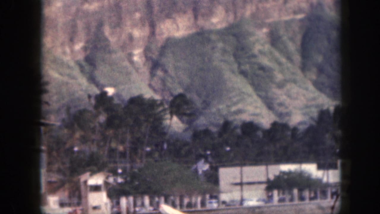 1959 Hawaii shoreline frame with palms, low structures, and steep mountain slopes in faded 8mm color.