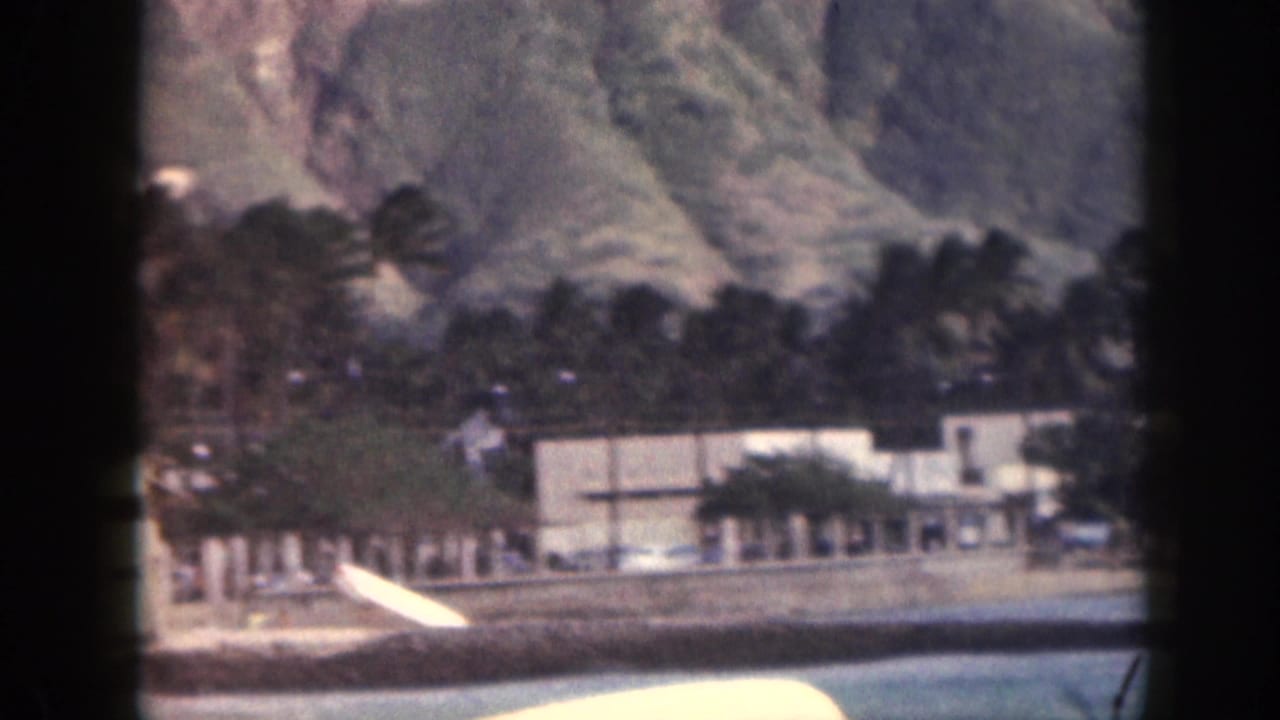 1959 Hawaii home-movie frame showing an outrigger canoe on turquoise water below palms, low buildings, and mountains.