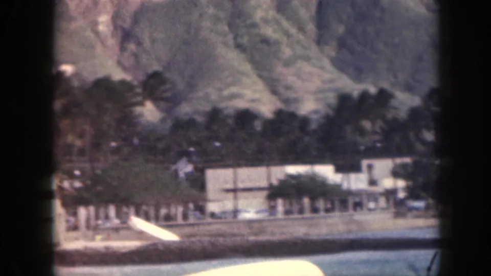 1959 Hawaii home-movie frame showing an outrigger canoe on turquoise water below palms, low buildings, and mountains.