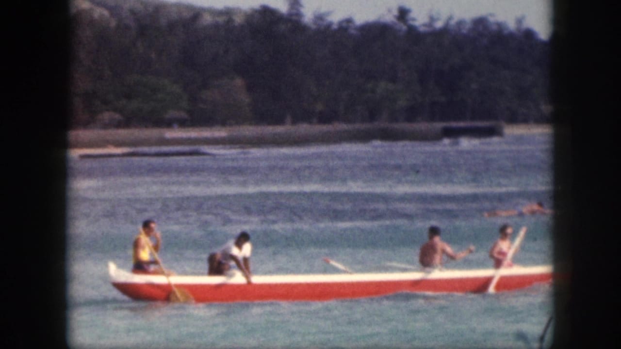 1959 Hawaii home-movie frame showing four people paddling a red-and-white outrigger canoe across blue coastal water.