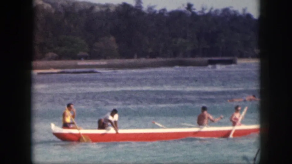 1959 Hawaii home-movie frame showing four people paddling a red-and-white outrigger canoe across blue coastal water.