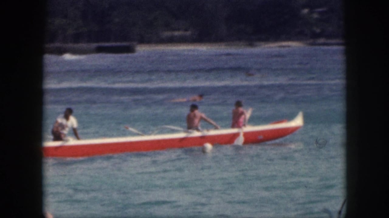 Outrigger canoe with paddlers crossing shallow water, with swimmers and shoreline behind.