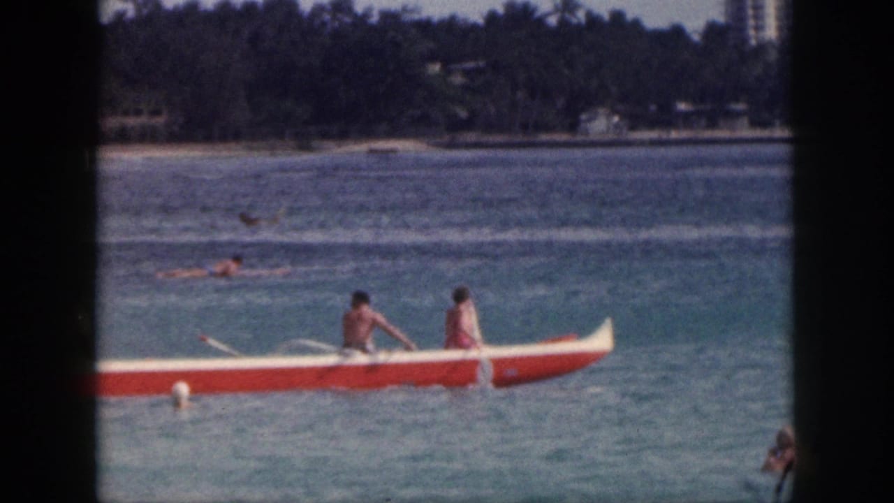 1959 Hawaii home-movie view of an outrigger canoe passing through blue water with swimmers and a distant shoreline.