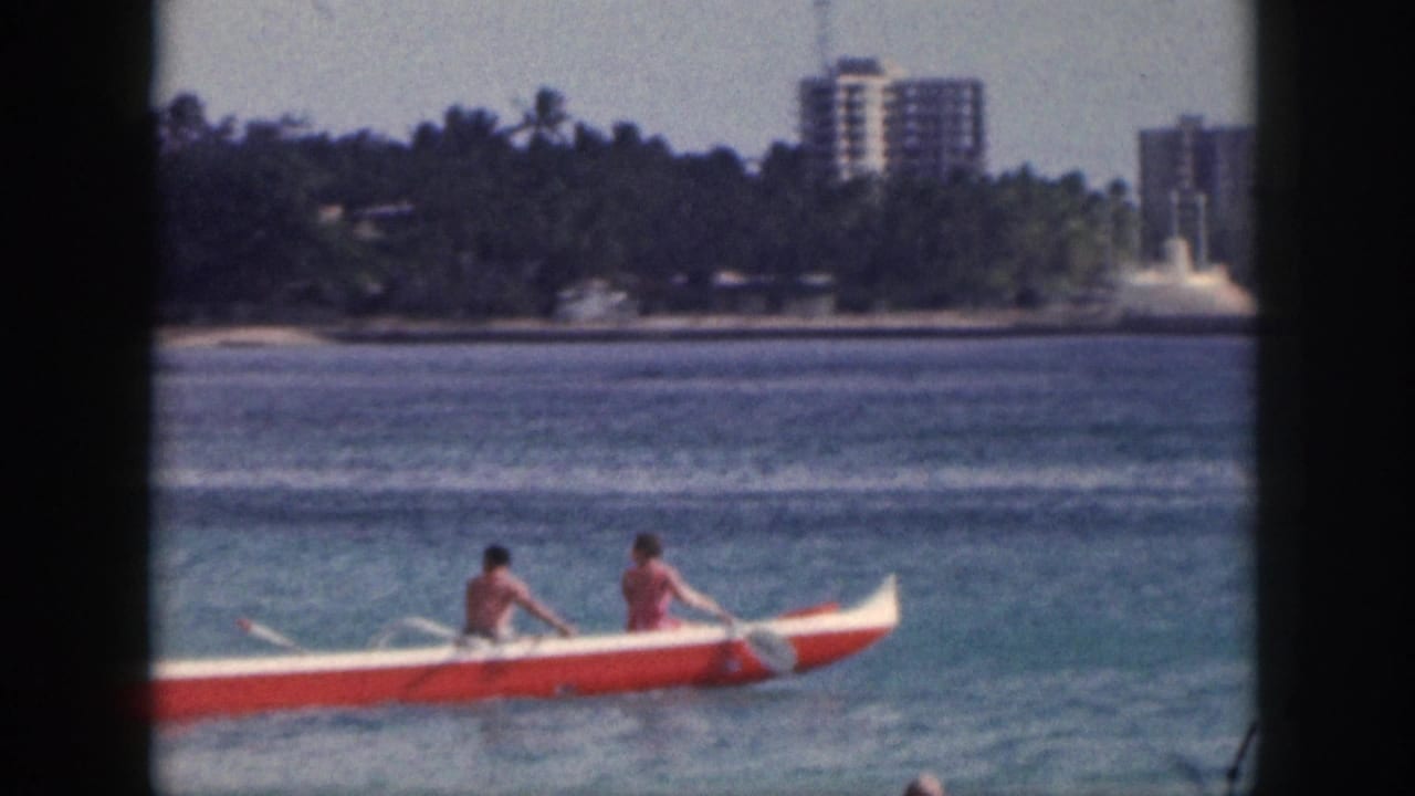 1959 Hawaii frame showing an outrigger canoe on coastal water, with trees and distant high-rise buildings along the shore.