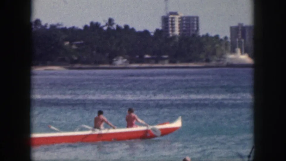 1959 Hawaii frame showing an outrigger canoe on coastal water, with trees and distant high-rise buildings along the shore.
