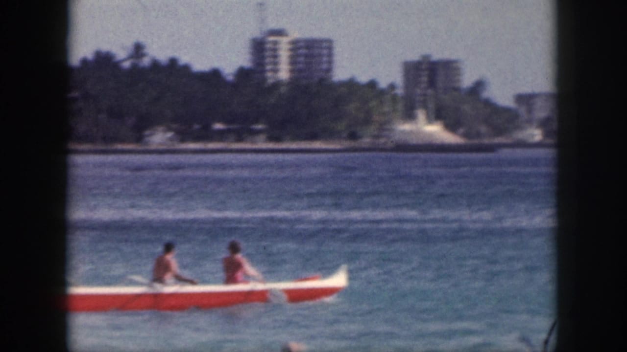1959 Hawaii 8mm frame showing two people in a red and white outrigger canoe near a palm-lined coast and distant high-rise buildings.
