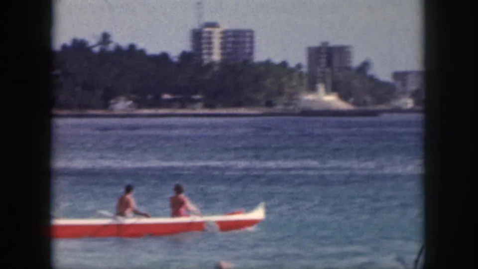 1959 Hawaii 8mm frame showing two people in a red and white outrigger canoe near a palm-lined coast and distant high-rise buildings.