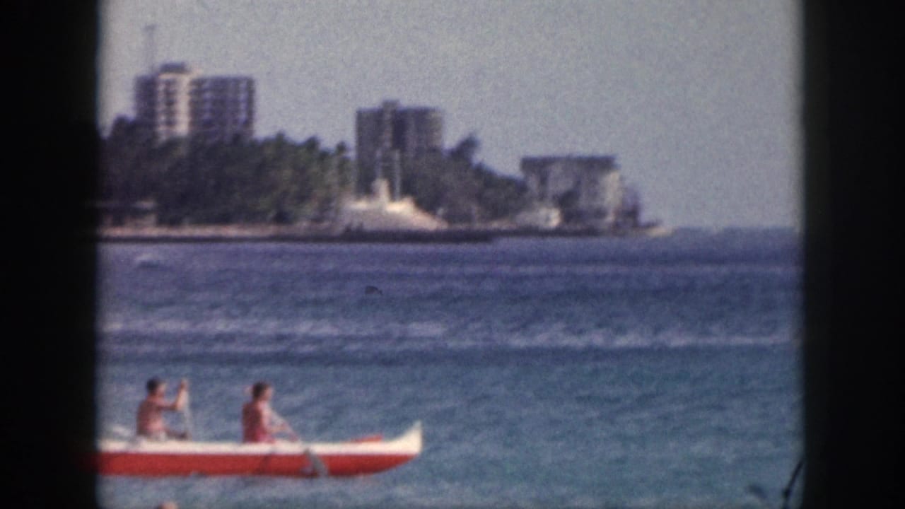 A red and white outrigger canoe with two paddlers crosses the water with shoreline buildings in the background.