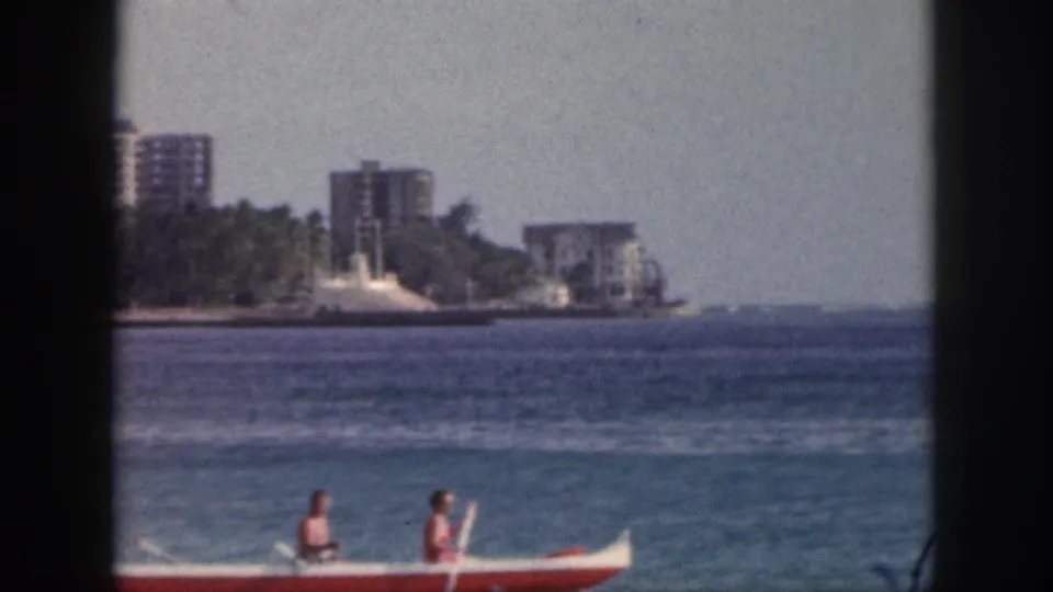 1959 Hawaii 8mm frame showing the canoe near the bottom of the picture, with blue water, palms, and coastal buildings beyond.