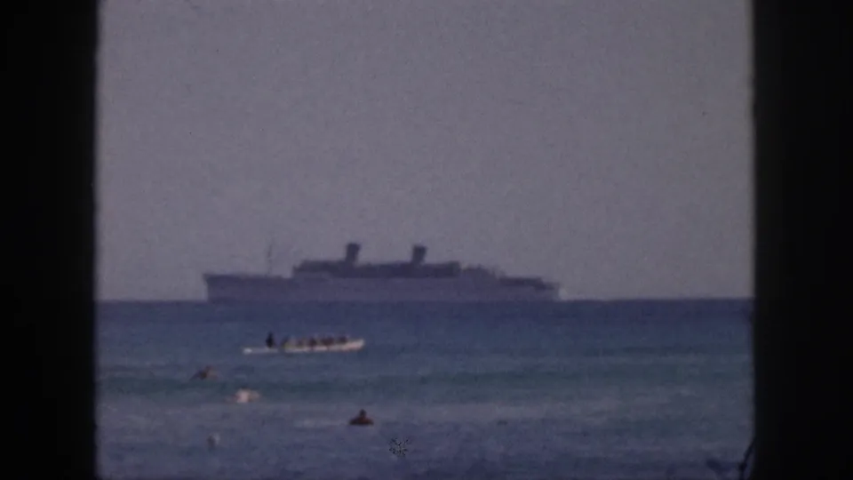 1959 Hawaii 8mm frame showing a small boat carrying people in the foreground with a larger ship farther out at sea.