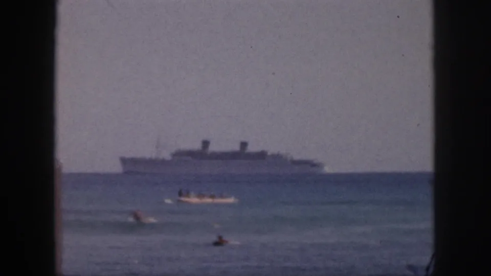 1959 Hawaii beach footage shows swimmers and a small motorboat in the foreground, with a large distant ship under a hazy sky.