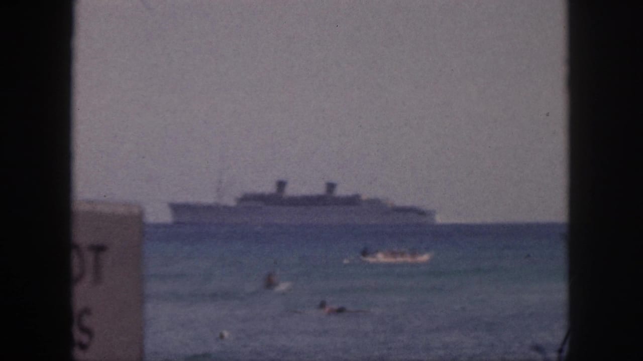 The Hawaii shoreline view includes a large ship offshore, a small boat, swimmers, soft surf, and part of a sign at the frame edge.