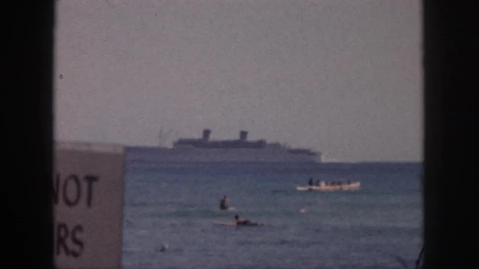A large distant ship remains near the horizon as swimmers and a small boat move across the water, with partial sign lettering at left.