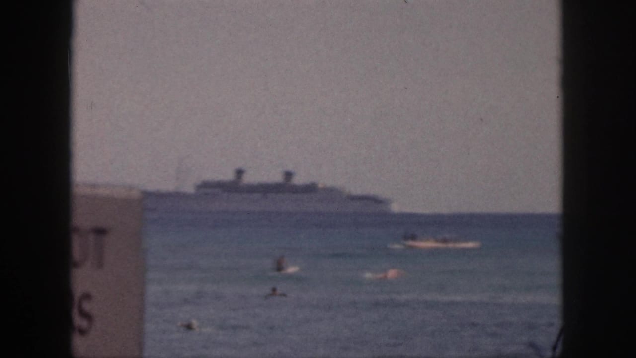Swimmers in ocean water with a large ship offshore and a partial sign at the left edge.