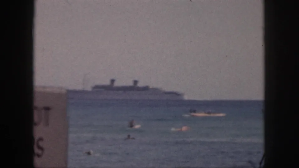 1959 Hawaii 8mm frame showing swimmers near shore, a large ocean liner in the distance, and a cropped beach sign at the left edge.