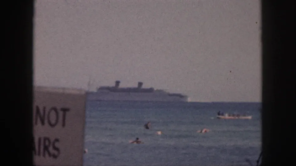 1959 Hawaii beach view with swimmers in the water, a distant passenger ship, a small boat, and a partly readable sign near the center-left.