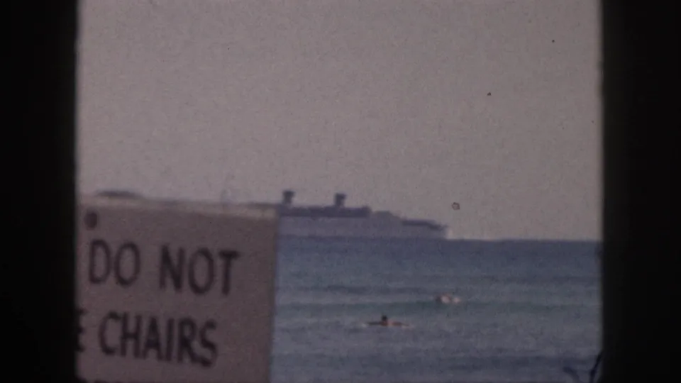 1959 Hawaii beach frame showing a cropped 'DO NOT' and 'CHAIRS' sign in front of swimmers and a distant passenger ship.