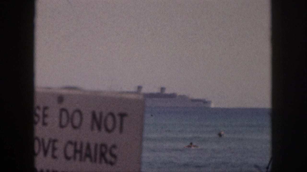 Distant ocean liner beyond swimmers and a foreground sign warning not to move chairs.